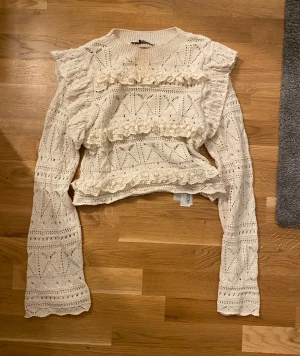Vit spetsblus med volanger från Zara - Superfin vit blus från Zara med långärmade ärmar och detaljerade spetsvolanger framtill. Blusen har ett stickat mönster och är dekorerad med broderade blommor på volangerna. Perfekt för dig som gillar romantisk och feminin stil.