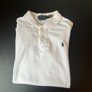 Vit pikétröja från Ralph Lauren - Vit pikétröja från Polo Ralph Lauren i slim fit-modell. Klassisk krage, två knappar och den ikoniska broderade loggan i mörkblått på bröstet. Tillverkad i bomull för en skön och stilren känsla.