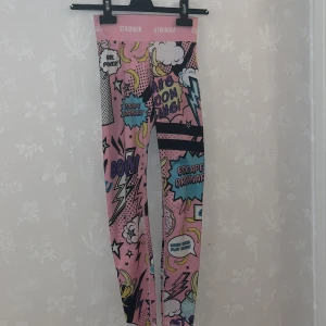 Rosa Stronger leggings med pop art-mönster - Färgglada leggings från Stronger i rosa med pop art-inspirerat tryck och text. Materialet är 85% polyester och 15% elastan, vilket gör dem stretchiga och bekväma för träning. Knappt använda (ungefär 1-2 gånger), inget hål eller liknande. Säljer pga för små.