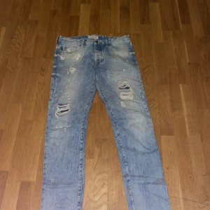 Ljusblå distressed jeans Scotch & Soda - Säljer ett par ljusblå jeans från Scotch & Soda med slitna detaljer och hål på benen för en riktigt cool look. Jeansen har klassisk femficksdesign och är tillverkade i bomull. Perfekta för dig som gillar en avslappnad och trendig stil.