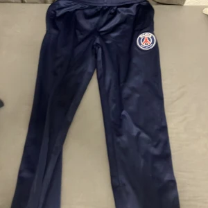 PSG träningsbyxor i mörkblått - Mörkblå träningsbyxor från PSG med klubbens logga på låret. Byxorna har elastisk midja och dragkedjor vid bensluten. Perfekta för fotbollsträning eller chill. Tillverkade i glansig polyester som ger en sportig look.