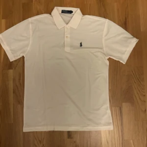 Vit pikétröja från Polo Ralph Lauren - Klassisk vit pikétröja från Polo Ralph Lauren med korta ärmar, krage och två knappar framtill. Tröjan har den ikoniska broderade loggan i mörkblått på bröstet och är tillverkad i mjuk bomull. Perfekt för en clean och stilren look.