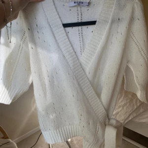 Vit omlottkofta från NA-KD XS - Superfin vit stickad kofta från NA-KD i omlottmodell med knytband i midjan. Koftan har ett hålmönster, bred ribbad kant och trekvartsärmar. Perfekt för lager-på-lager och ger en mysig men ändå trendig look. Storlek xs men passar också s