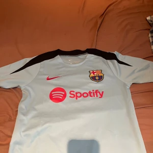 Barça Nike Dri-Fit fotbollströja M - Snygg ljusblå FC Barcelona fotbollströja från Nike med svarta detaljer på axlarna och röd Nike-logga samt Spotify-tryck på bröstet. Tröjan är tillverkad i Dri-Fit material som andas och har klubbmärket broderat på vänster bröst.