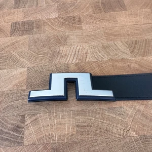 Svart skärp från J.Lindeberg i skinn - Snyggt svart bälte från J.Lindeberg i äkta skinn, tillverkat i Italien. Bältet har en unik, silverfärgad spänne i geometrisk form som verkligen sticker ut. Perfekt accessoar för att lyfta din outfit med en stilren och modern touch.