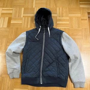 Quiltad bomberjacka från Indigo XL - Quiltad bomberjacka från Indigo i storlek XL. Jackan har marinblå kropp med grå ärmar, huva med snörning och dragkedja framtill. Ärmarna är mjuka och ribbade vid muddar, medan kroppen är lätt vadderad och mönstrad i rutor. 