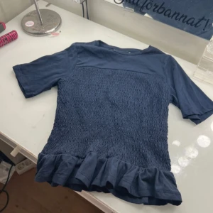 Mörkblå topp med volang från Gina Tricot - Supersöt mörkblå topp från Gina Tricot i bomull med korta ärmar, smockad midja och volangkant nedtill. Perfekt för dig som gillar en enkel men ändå detaljrik stil. T-shirten har en klassisk rund halsringning och är riktigt bekväm.