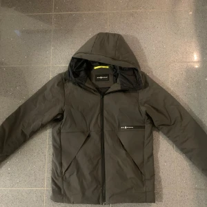 Mörk grå pufferjacka från Sail Racing - Säljer en mörk grå pufferjacka från Sail Racing med huva och dragkedja framtill. Jackan har två stora fickor, diskret logga på bröstet och. Perfekt för kalla dagar och har en clean, sportig look. Priset kan diskuteras vid snabb affär!! Hör av er vid minsta lilla fundering!!!!!!