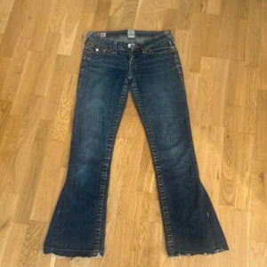 True Religion bootcut jeans blå, stl 26 - Snygga blå bootcut jeans från True Religion med klassiska kontrastsömmar. Jeansen har låg midja och ikoniska bakfickor med lock och knapp. Den jag köpte de av hade klippt vid benen därför de ser ut som de gör. Benlängd är 92 från midjan och Midje mått tvärs över är 37! Jeansen hade också ett hål vid fickan som visas på bild.
