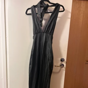 Svart långklänning med knyt i nacken - Snygg svart långklänning med djup v-ringning och knyt i nacken. Klänningen har en slät, glansig yta som ger en cool look och är ärmlös med en figurnära passform. Perfekt för dig som vill sticka ut med en stilren och elegant vibe.