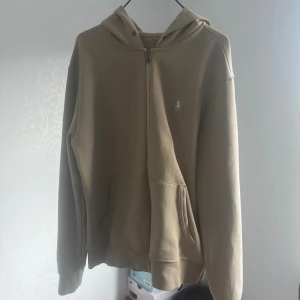 Beige hoodie från Polo Ralph Lauren - Snygg beige hoodie från Polo Ralph Lauren med klassisk logga broderad i vitt på bröstet. Tröjan har huva, dragkedja framtill och en stor känguruficka. Tillverkad i mjuk bomull och har långa ärmar. Perfekt för chill dagar!