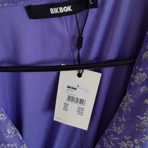 Lila blommig kortklänning Bik Bok Storlek:L - Superfin lila kortklänning från Bik Bok med blommigt mönster och volangdetaljer. Storlek:L. Klänningen har omlottknytning i midjan och är tillverkad i ett lätt, transparent syntetmaterial med korta ärmar. Perfekt för dig som vill ha en söt och trendig look. Prislappen är kvar.