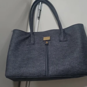 Mörkgrå handväska från Ninoel - Snygg mörkgrå handväska från Ninoel med subtilt fiskbensmönster. Väskan har två rejäla handtag, guldfärgade detaljer och dragkedja upptill. Perfekt storlek för att få plats med allt du behöver. Materialet känns slitstarkt och har en elegant struktur.