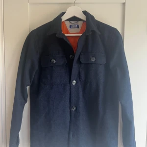 Mörkblå overshirt från Jack & Jones - Snygg mörkblå overshirt från Jack & Jones med orange quiltat foder. Jackan har två bröstfickor med knappar och klassisk krage. Perfekt för lager-på-lager och har en stilren look med raka linjer och lång ärm.