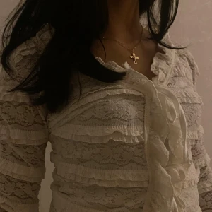 Vit spetsblus med volanger och knyt - Supersöt vit blus i spets med långa ärmar, volangdetaljer och knyt framtill. Blusen har transparent spets och romantisk känsla, perfekt för dig som gillar en feminin och trendig stil. Sitter snyggt figurnära och har v-ringad urringning.