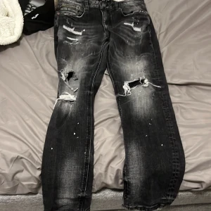 Svarta slitna jeans med hål - Svarta jeans med coola slitningar och hål på knäna, samt vita färgstänk för en edgy look. Jeansen har en rak passform och tydliga slitdetaljer framtill. Perfekta för dig som gillar streetstyle och vill sticka ut.