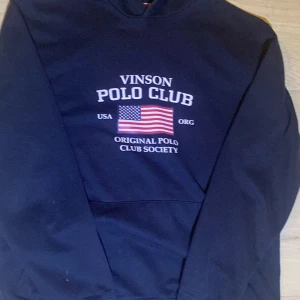 Mörkblå hoodie Vinson Polo Club - Mörkblå hoodie från Vinson Polo Club med tryck framtill och amerikansk flagga. Klassisk känguruficka och huva. Perfekt för dig som gillar sportig och avslappnad stil.