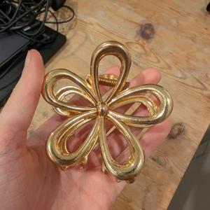 Guldig blomformad hårklämma - En stor hårklämma i metall med glansig guldfinish, formad som en stiliserad blomma med fem breda kronblad. Perfekt accessoar för att lyfta din frisyr med en trendig och iögonfallande detalj.