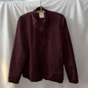 Vinröd overshirt från Wood Wood - Snygg vinröd overshirt från Wood Wood med broderad W-logga på bröstet. Jackan har klassisk krage, knappar framtill och två sidofickor. Materialet är glansigt och känns lätt, perfekt för lager-på-lager. Passformen är normal och ärmarna är långa.