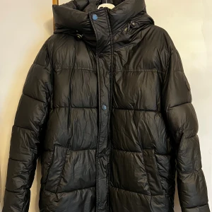 Svart pufferjacka från Indigo Jeanscode - Svart pufferjacka från Indigo Jeanscode med hög krage, huva och broderad logga på ärmen. Jackan har dragkedja med gul textdetalj, stora fickor och quiltad design. Yttermaterialet är i polyester och insidan är fodrad med mjuk fleece för extra värme.