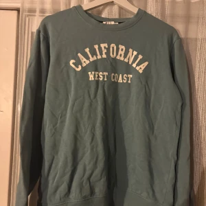 Blå sweatshirt California KappAhl - Snygg blå sweatshirt från KappAhl med trycket 'California West Coast' i vitt framtill. Tröjan har rund halsringning, långa ärmar och ribbade muddar vid ärmslut och nederkant. Perfekt för en avslappnad och cool stil.