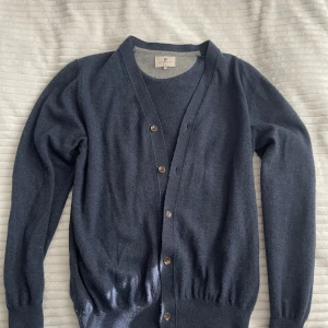 Hansen & Jacob kofta - Mörkblå kofta från Hansen & Jacob i mjuk merinoull med knappar framtill och snygga mockadetaljer på armbågarna. Tröjan är knappt använd och nypriset ligger på straxt över 2000kr