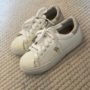 Vita sneakers från Tommy Hilfiger - Snygga vita sneakers från Tommy Hilfiger med guldfärgad logga på sidan och dragkedja vid snörningen. Skorna är i skinn med rund tå och platt sula. Perfekta för dig som gillar klassisk och stilren look med en twist. Skorna är knappt använda. Priset kan diskuteras.