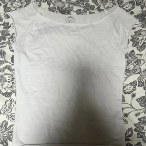 Vit topp från Urban Outfitters - En enkel och stilren vit topp från Urban Outfitters. Modellen har korta ärmar och en bred, rundad halsringning. Tillverkad i mjuk bomull som känns skön mot huden. Perfekt att matcha med jeans eller kjol för en clean look.