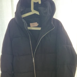 Svart pufferjacka med huva från Nelly - Svart pufferjacka från Nelly i storlek 38. Jackan har en stor huva, dragkedja framtill och bälte i midjan för extra snygg passform. Perfekt för kalla dagar när du vill hålla dig varm och ändå se cool ut.