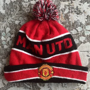 Manchester United mössa från New Era - Röd och svart Manchester United mössa med vitrandiga detaljer och stor tofs på toppen. Broderad klubbmärke framtill och texten 'MAN UTD' runt om. Tillverkad i mjukt syntetmaterial, perfekt för kalla dagar på läktaren.