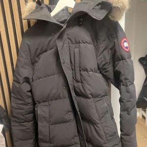 Svart Canada Goose Carson Parka M - Säljer en svart Canada Goose Carson Parka med pufferdesign och huva med äkta päls. Jackan har klassisk patch på ärmen, flera fickor och dragkedja med knappslå. Perfekt för kalla dagar och riktigt snygg vinterstil.