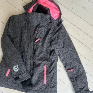 Leopardmönstrad vinterjacka Woxo 158 - Grym vinter/skid från Woxo by KappAhl i leopardmönster. Yttertyget är mörkgrått med svarta fläckar och detaljer i rosa, som dragkedjor och foder. Jackan har huva, flera fickor och quiltat rosa innerfoder. Perfekt för kalla dagar och snygg streetstil.