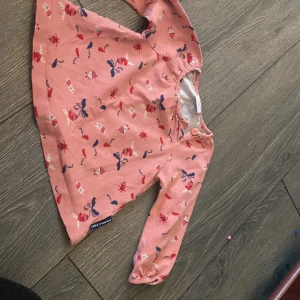 Rosa blus med djurmönster Polarn O. Pyret - Supersöt rosa blus från Polarn O. Pyret med lekfullt mönster av små djur i rött, vitt och blått. Blusen har långa ärmar med resår vid handlederna och knäppning vid axeln för enkel påklädning. Perfekt för vår och sommar!