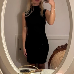 Svart ärmlös kortklänning - Snygg svart kortklänning med hög hals och ärmlös design. Klänningen har en tight passform som framhäver figuren och är perfekt för dig som gillar en stilren och klassisk look. Enkel att styla med accessoarer för en personlig touch.