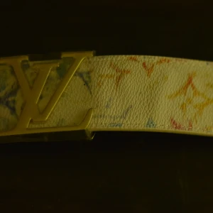 Louise Vuitton vitt bälte med guld spänne - Snyggt vitt bälte från Louise Vuitton med ikoniskt spänne i guld. Bältet har färgglada monogramdetaljer i blått, rött och gult på präglat skinn. Insidan är ljusbrun och bältet känns lyxigt och trendigt – perfekt för att lyfta din outfit.