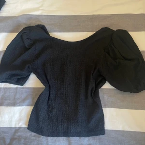 Svart blus med puffärmar - Säljer en svart blus med snygg struktur och stora puffärmar. Toppen har en lätt croppad passform och bred urringning som ger en trendig siluett. Perfekt att styla med jeans eller kjol för en cool look.