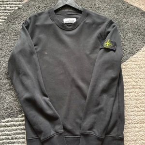 Svart sweatshirt från Stone Island - Svart långärmad sweatshirt från Stone Island med klassisk rund hals och ikonisk patch på vänster ärm. Tröjan är tillverkad i mjuk bomull och har ribbade muddar vid ärmslut och nederkant. Perfekt för en clean och stilren look.