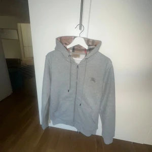 Grå Burberry hoodie med rutig huva - Grå hoodie från Burberry med klassisk rutig insida i huvan och längs dragkedjan. Tröjan har huva med snörning, två fickor fram och subtilt Burberry-logo broderat på bröstet. Tillverkad i mjuk bomull och polyesterblandning, perfekt för chill dagar. Används ett par gånger, pris kan diskuteras!