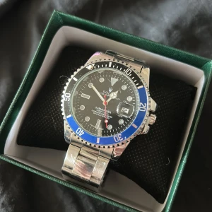 Submariner med blå och svart bezel - Säljer en stilren Submariner med blå och svart bezel, svart urtavla och datumvisning. Klockan har en robust länk i silverfärgat stål och tydliga indexmarkeringar. Perfekt för dig som gillar klassisk design med en modern touch. Levereras i original ask.