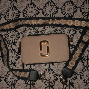 Beige axelväska Marc Jacobs med bred rem - Snygg beige axelväska från Marc Jacobs med guldig logga framtill och bred, justerbar rem med text. Väskan har svarta detaljer och dragkedja i guld. Materialet är skinn och väskan har ett extra fack på baksidan. Perfekt för dig som vill ha en trendig och praktisk väska.