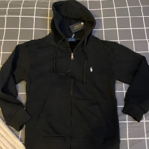 Svart hoodie från Polo Ralph Lauren - Svart hoodie från Polo Ralph Lauren med dragkedja framtill, snörning i huvan och klassisk broderad logga på bröstet. Mjuk bomullskvalitet, känguruficka och ribbade muddar. Perfekt för en chill och stilren look.