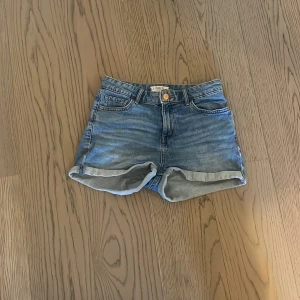 Blå jeansshorts från Lindex stl 158 - Snygga blå jeansshorts från Lindex med klassisk femficksdesign och uppvikta ben. Shortsen har justerbar midja med knappresår men ena av de ör sönder. Perfekta för sommarens alla häng och chill dagar.                                                                        Pris går att diskutera!