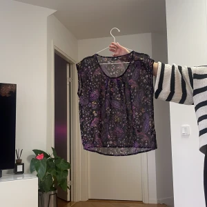 Svart blommig blus med lila paisley - Säljer en luftig svart blus från JC med blommigt och paisleymönster i lila och rosa toner. Blusen är ärmlös och har rund halsringning. Perfekt för dig som gillar transparenta och mönstrade toppar med en unik vibe.
