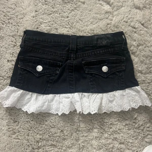 Svart jeanskjol True Religion - svart jeanskjol från True Religion med silverknappar och bakfickor. Kjolen har en vit broderad volangkant längst ner🥰