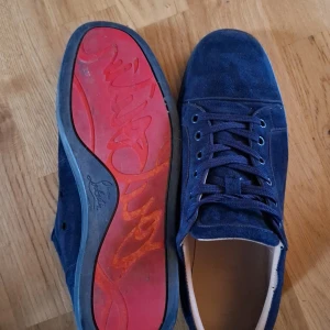 Blå mockasneakers från Christian Louboutin - Snygga sneakers i mörkblå mocka från Christian Louboutin med klassisk röd sula och diskret logga. Skorna har rund tå, platt sula och snörning. Insidan är i ljust skinn och utsidan i lyxig mocka. Perfekta för dig som vill sticka ut med exklusiva detaljer.