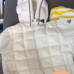 Vit Moncler pufferjacka med huva - Snygg vit pufferjacka från Moncler med quiltad framsida, stickade ärmar och stor vadderad huva med logga och rödblå detaljer. Jackan har dragkedja framtill, ribbade muddar och Moncler-märke på ärmen. Perfekt för kyliga dagar när du vill vara både varm och stilren.