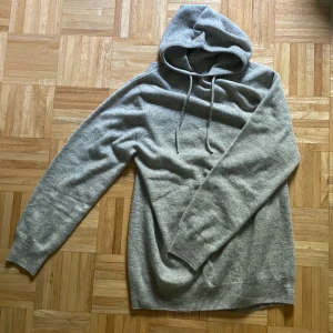 Grå hoodie i mjuk stickad ull - Säljer en stilren grå hoodie i mjuk stickad ull. Tröjan har huva med snörning och långa ärmar. Perfekt för dig som gillar en enkel och clean look. Passformen är normal och modellen är klassisk utan tryck eller mönster.