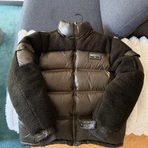 Brun pufferjacka från Christian Lane ( luva följer med oxå )  - Snygg brun pufferjacka från Christian Lane med teddy-detaljer på axlar och ärmar. Jackan har hög krage, dragkedja framtill och en badge på bröstet. Materialmix av polyester och teddy ger både värme och cool look. Perfekt för kalla dagar.