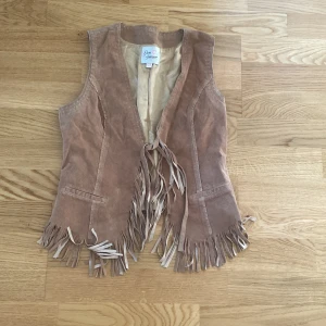 Beige mockaväst med fransar Love Forever - Säljer en beige mockaväst från Love Forever med snygga fransar längs nederkanten och framtill. Västen har två fickor och knytband framtill. Perfekt för dig som vill ha en cool westerninspirerad look.