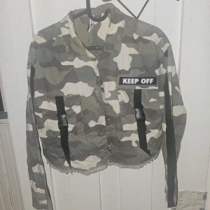 Camouflage overshirt från FB Sister - Snygg camouflagefärgad overshirt från FB Sister med coola svarta spänndetaljer och texten 'KEEP OFF' på bröstet. Jackan är croppad med rå kant nertill och har knappar framtill. Perfekt för dig som vill sticka ut med streetstyle.